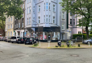 Eine Straßenecke. Im Hintergrund ein himmelblaues Mehrfamilienhaus, Altbau.