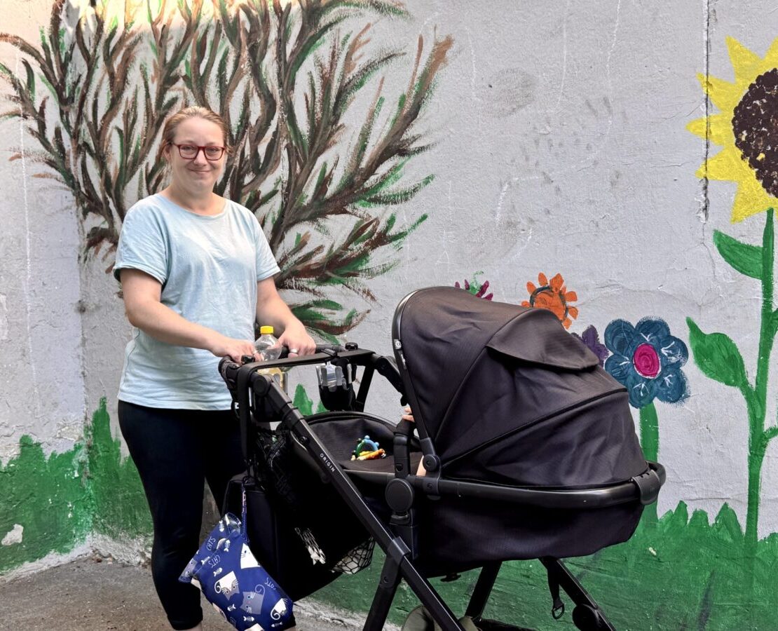 Auf dem Bild sieht man die Mutter Jenny, in die Kamera lächeln, vor sich hat sie einen schwarzen Kinderwagen in dem ihr Sohn drin ist, dessen sieht man nicht.