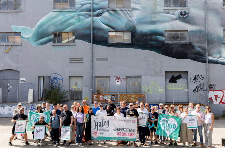 Auf dem Bild sieht man die verschiedene Akteure des Hafenspaziergangs 2026. Sie stehen. vor einem Gebäude und halten verschiedene Banner in die Luft.