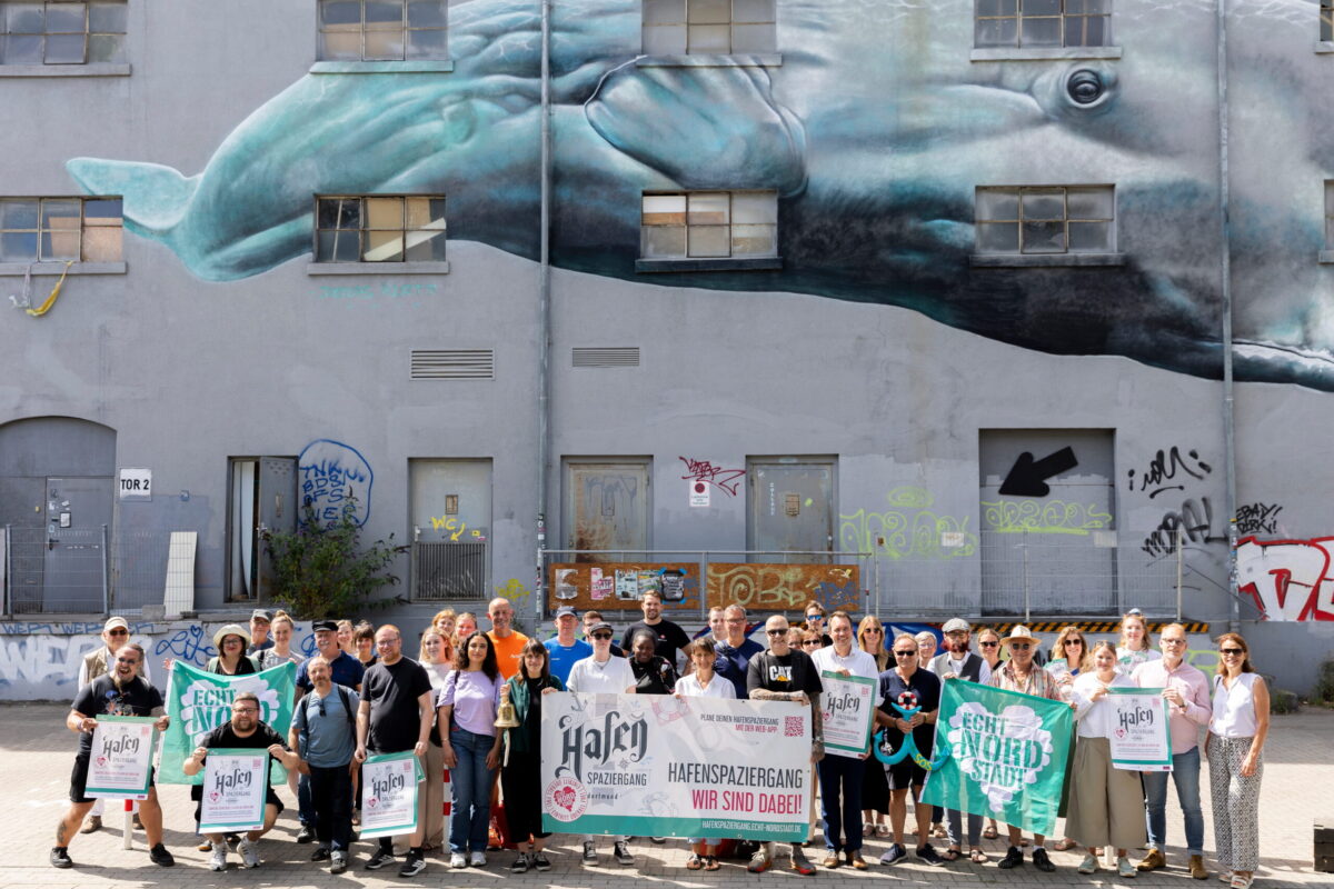 Auf dem Bild sieht man die verschiedene Akteure des Hafenspaziergangs 2026. Sie stehen. vor einem Gebäude und halten verschiedene Banner in die Luft.