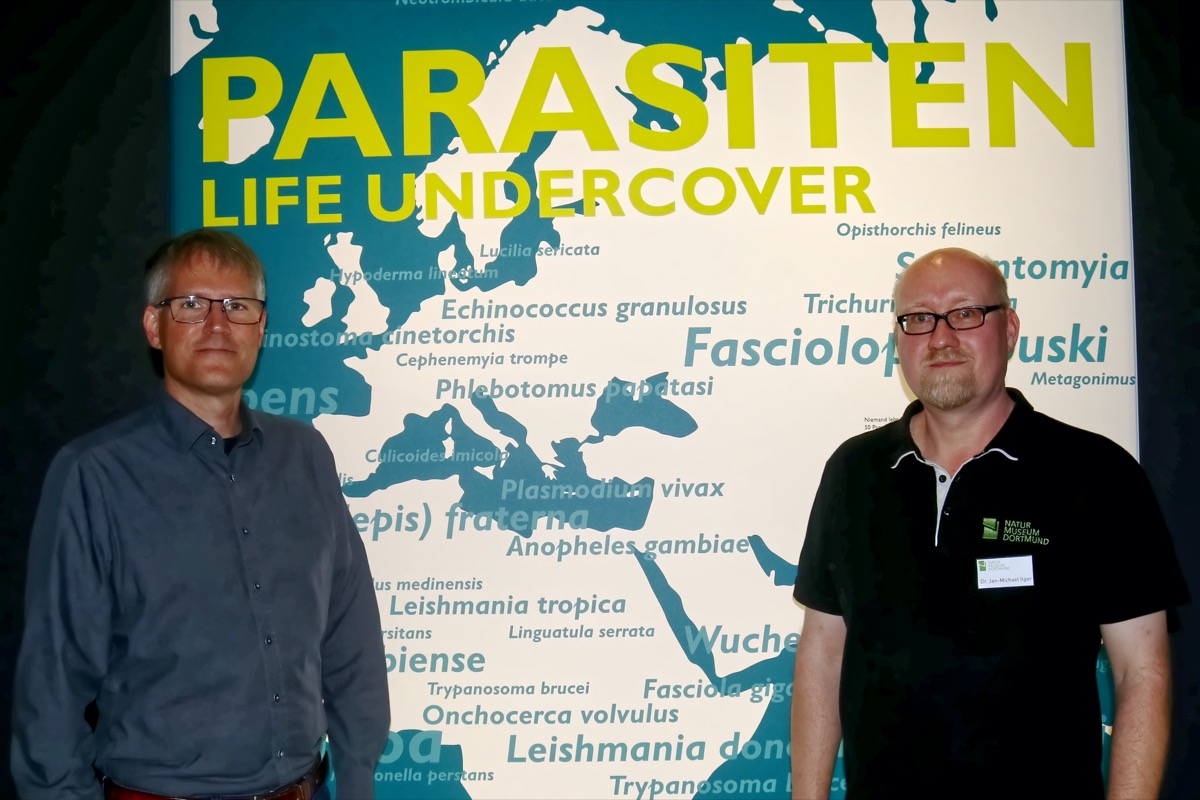 Zwei Männer vor einer Werbetafel mit der Aufschrift : Parasiten , Life Wundercover