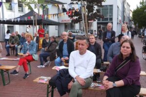 Menschen sitzen auf Biertischbänken und hören zu.
